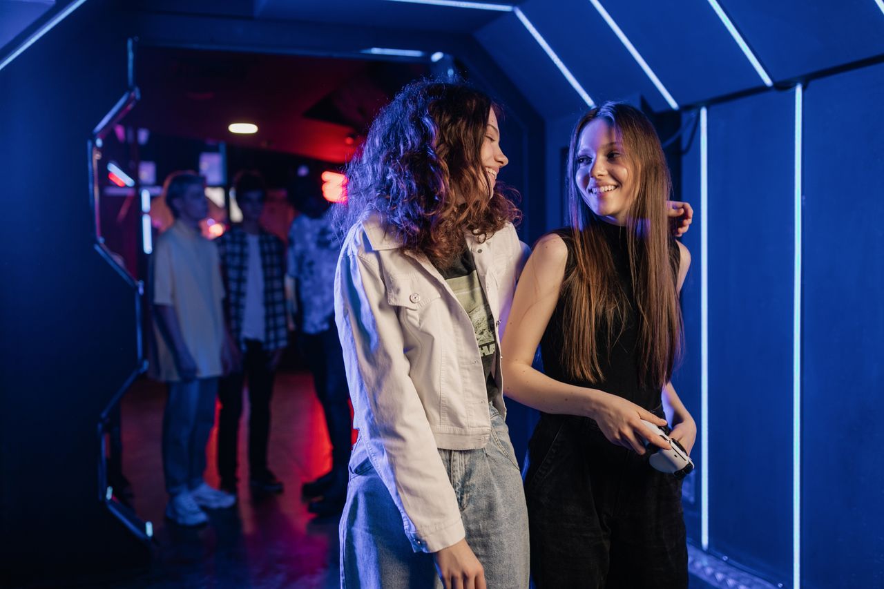 Two women laughing and gaming indoors in a neon-lit room, enjoying entertainment.